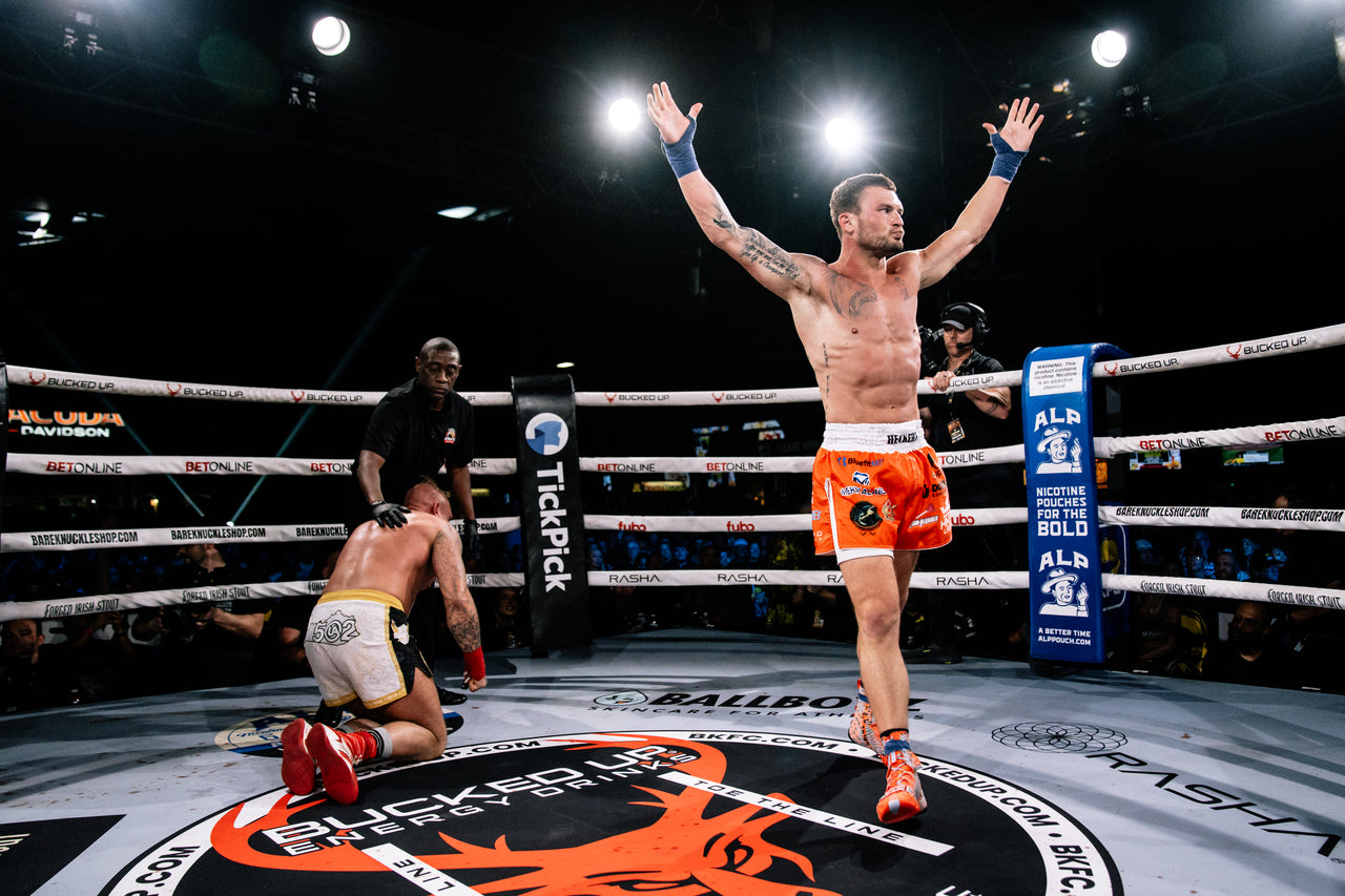 Boxer celebrating victory in a boxing ring with opponents on the ground.